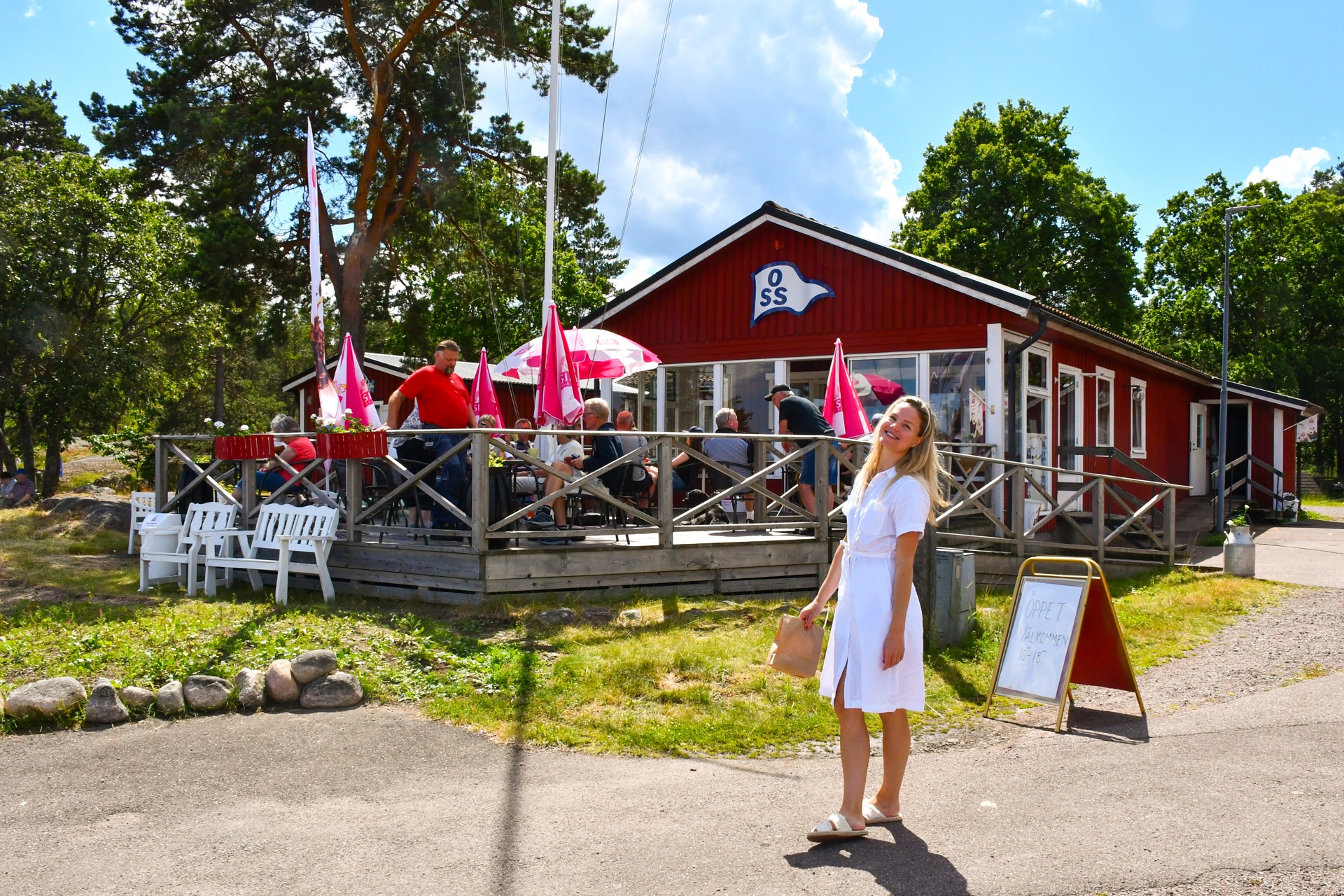 Glad kvinna med en papperspåse från Kafé Fridens framför den röda byggnaden med uteservering en sommardag.