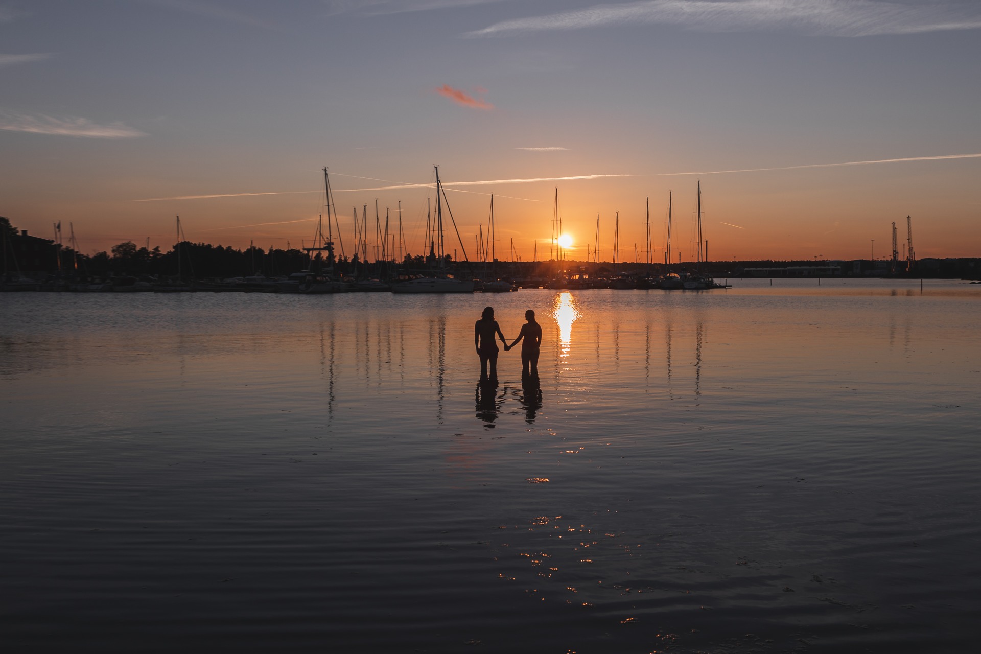 Bild tagen på två personer bakifrån som håller hand i havet med solnedgången framför sig och segelbåtar i Oskarshamn,