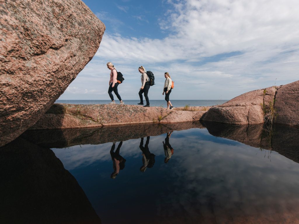 Tre personer som går på klippor på Blå jungfrun mot en större klippa med havet runt omkring.
