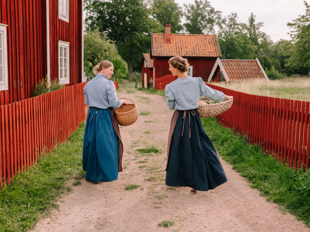 Två personer i 1900-talskläder som går på gården i kulturreservatet Stensjö by.