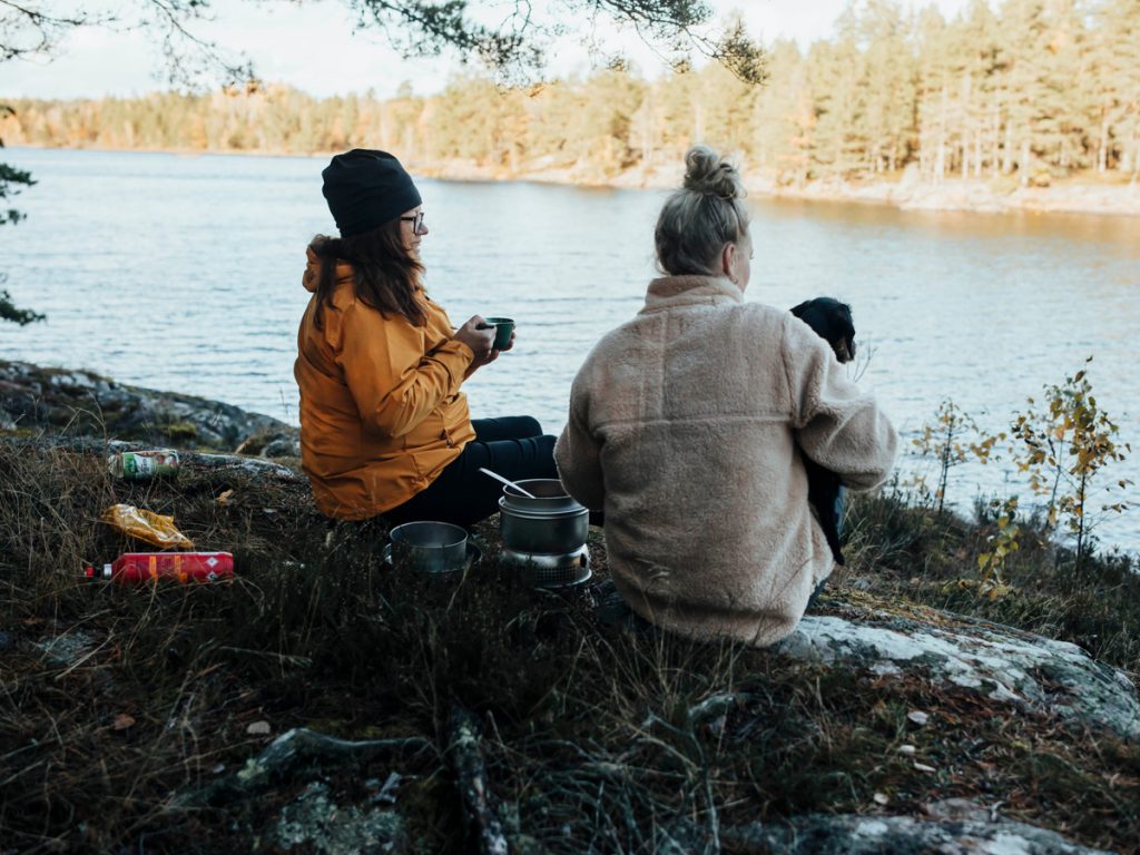 Två personer som sitter på en klippa och äter mat utomhus med utsikt över en sjö på Ostkustleden.