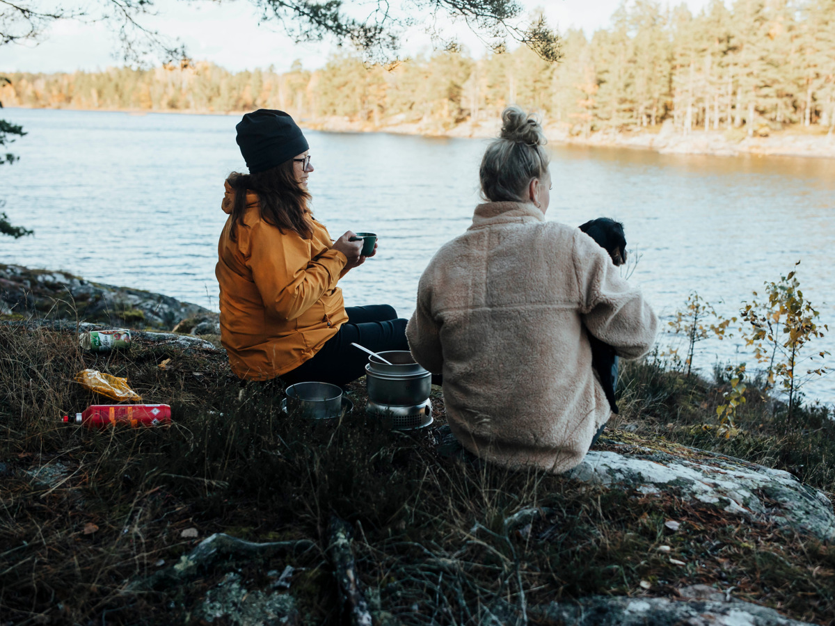 Två personer som sitter på en klippa och äter mat utomhus med utsikt över en sjö på Ostkustleden.