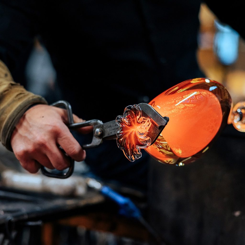 En glödhet glasbit som ett par händer håller fast i med hjälp av en svart tång.