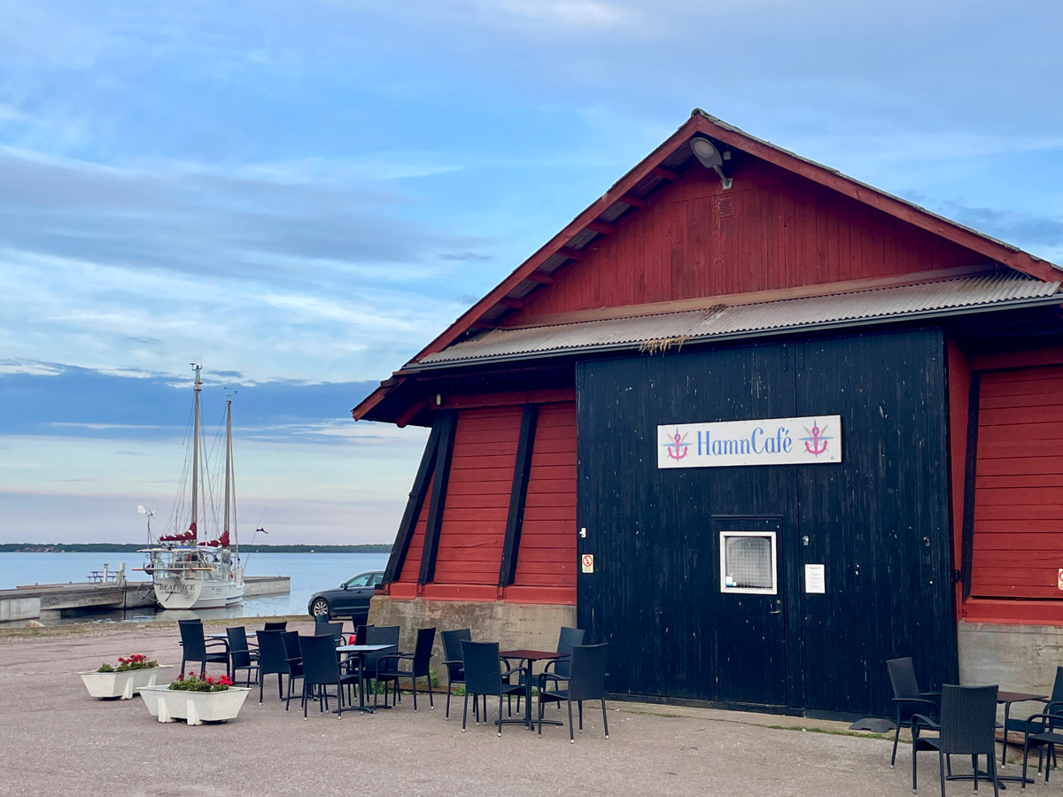 Bild på utsidan av Påskallaviks Hamncafé. En röd ladubyggnad med havet och segelbåtar i bakgrunden.