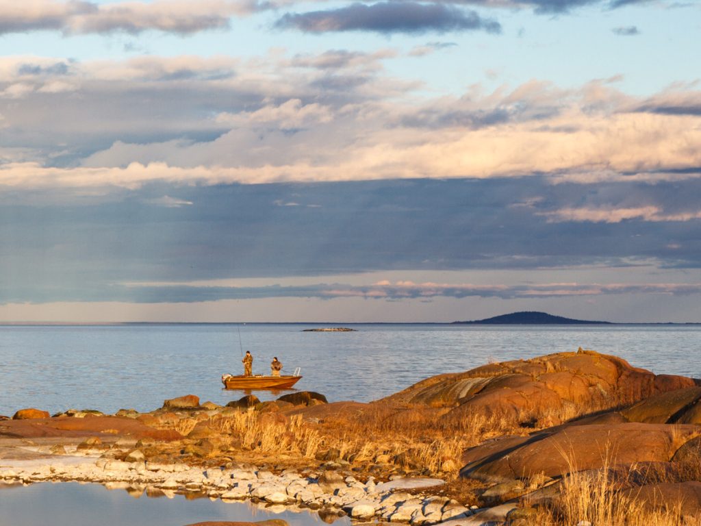 Bild på havet med en fiskebåt och två personer som fiskar med nationalparken Blå jungfrun i bakgrunden.