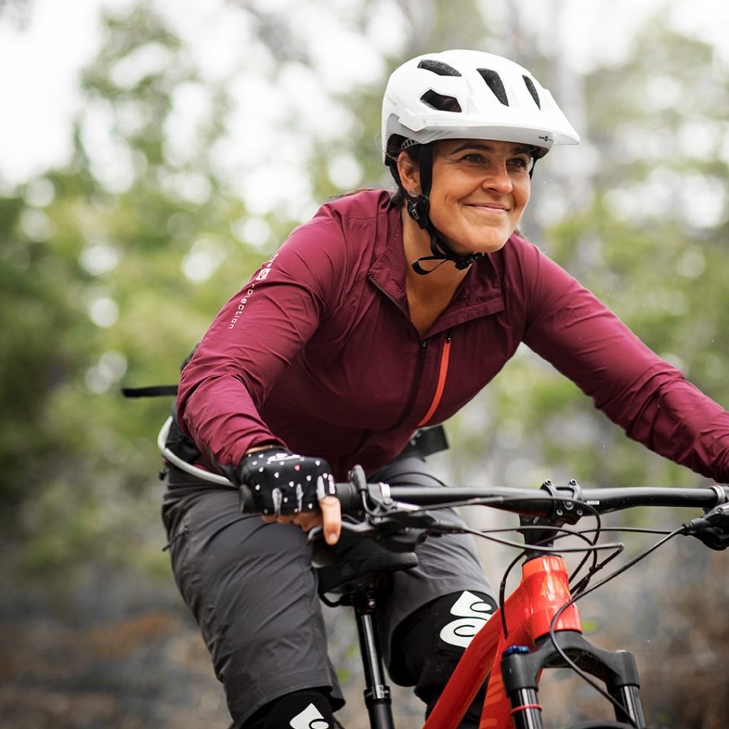 Kvinna i röd jacka och vit hjälm cyklar på en rödorange mountainbike genom skogen.