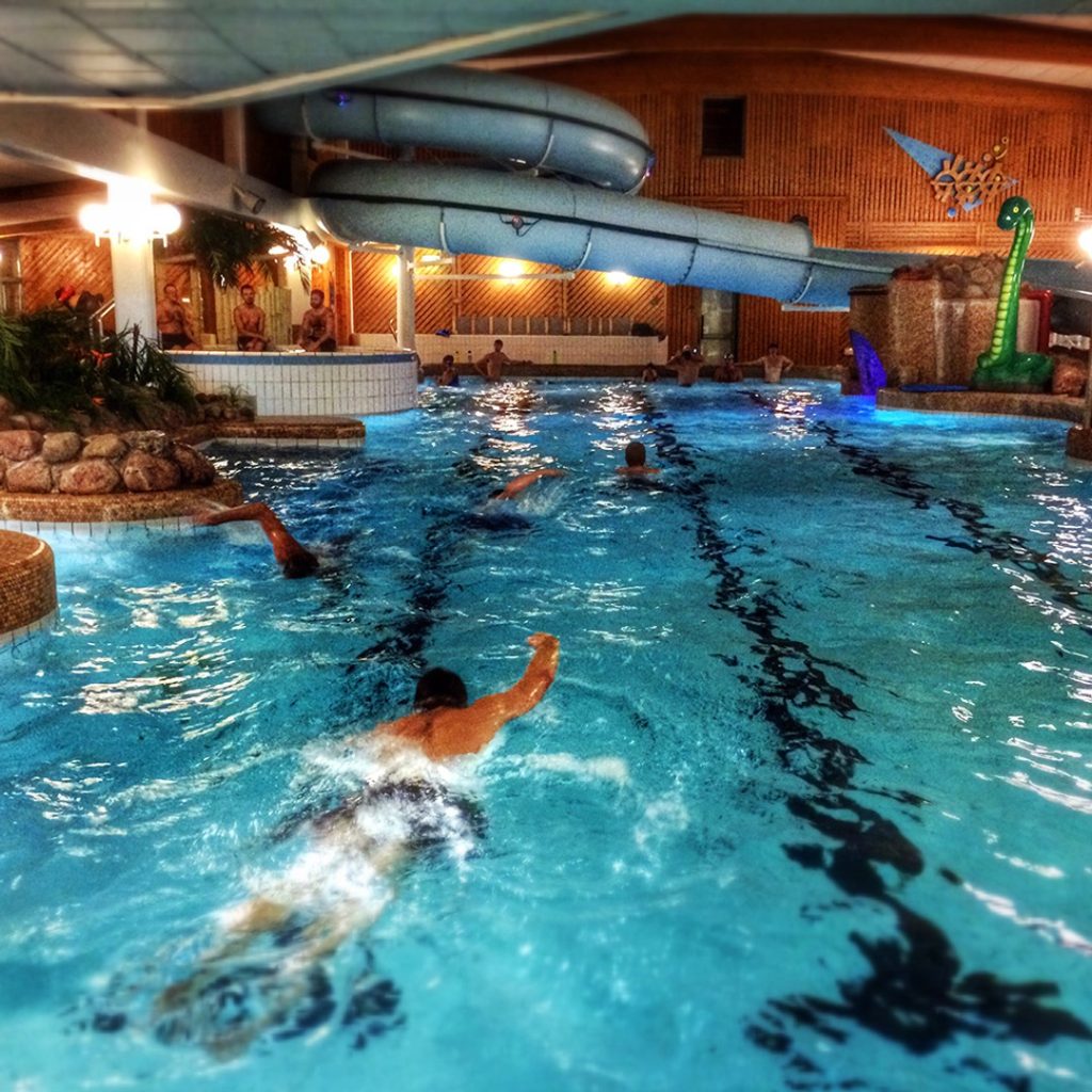 Flera personer som simmar crawl i en bassäng, ovanför vilken man kan skymta en vattenrutschbana.