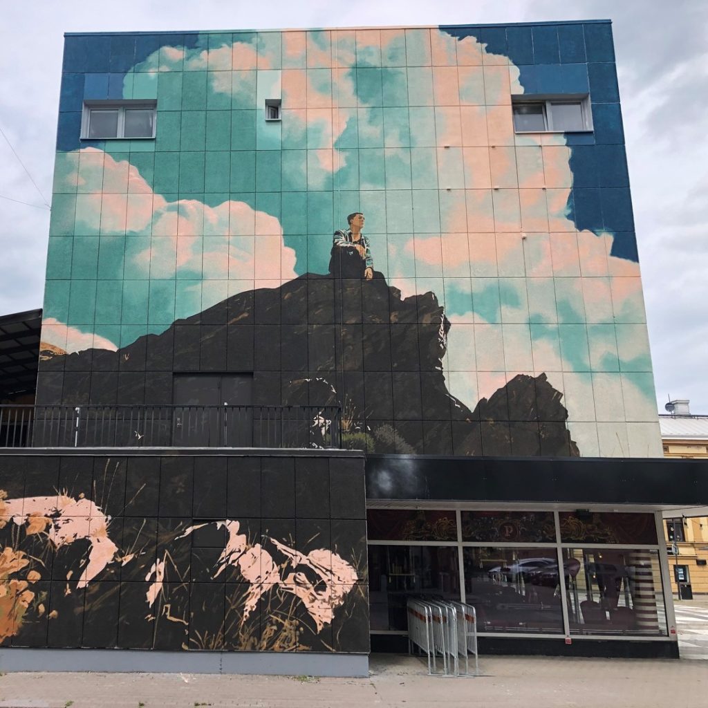 Väggmålning i centrala Oskarshamn föreställande en man blickandes ut från en hög klippa med blå himmel och vita moln i bakgrunden.