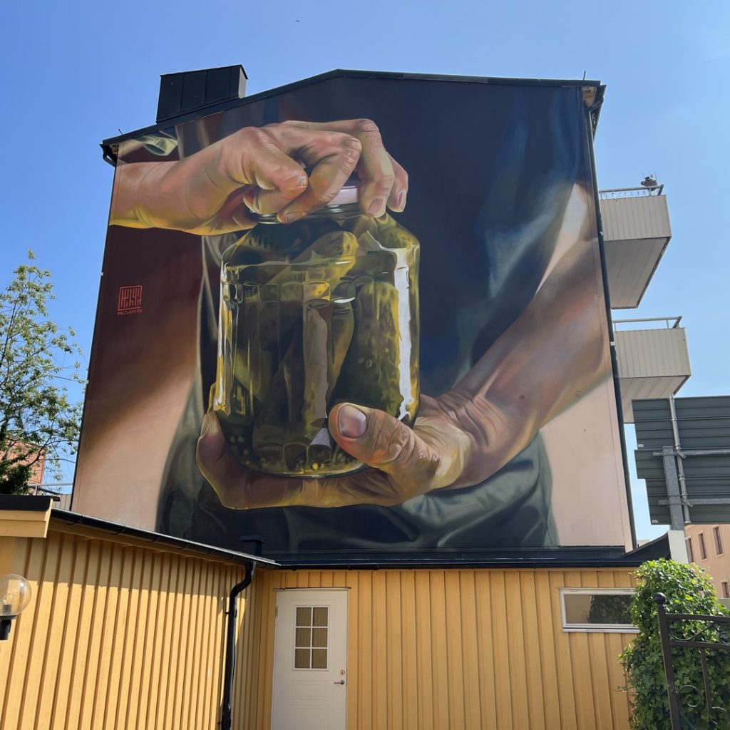 Väggmålning i centrala Oskarshamn föreställande två händer som försöker öppna en burk med inlagda gurkor, i vilken en människogestalt reflekteras.