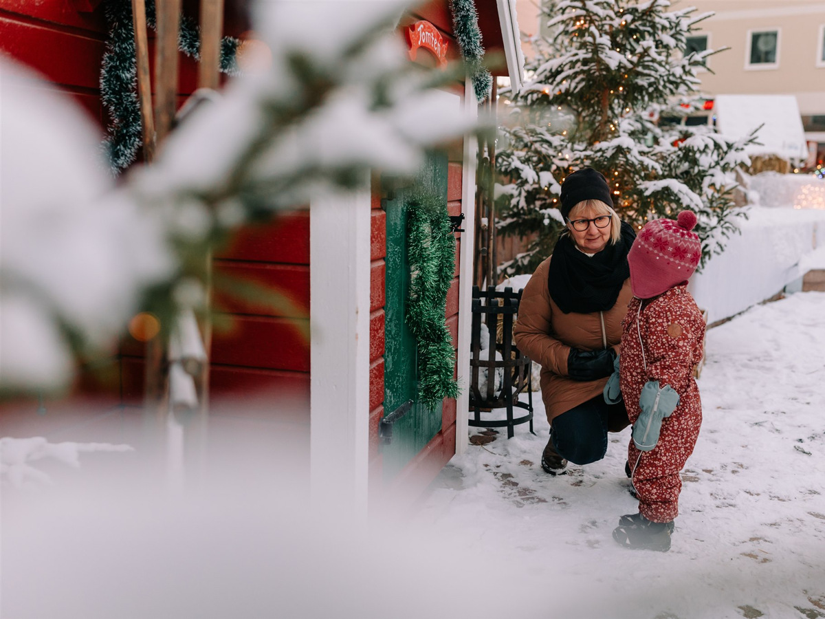 En farmor och ett barn vid en stuga på vintern med snö omkring.