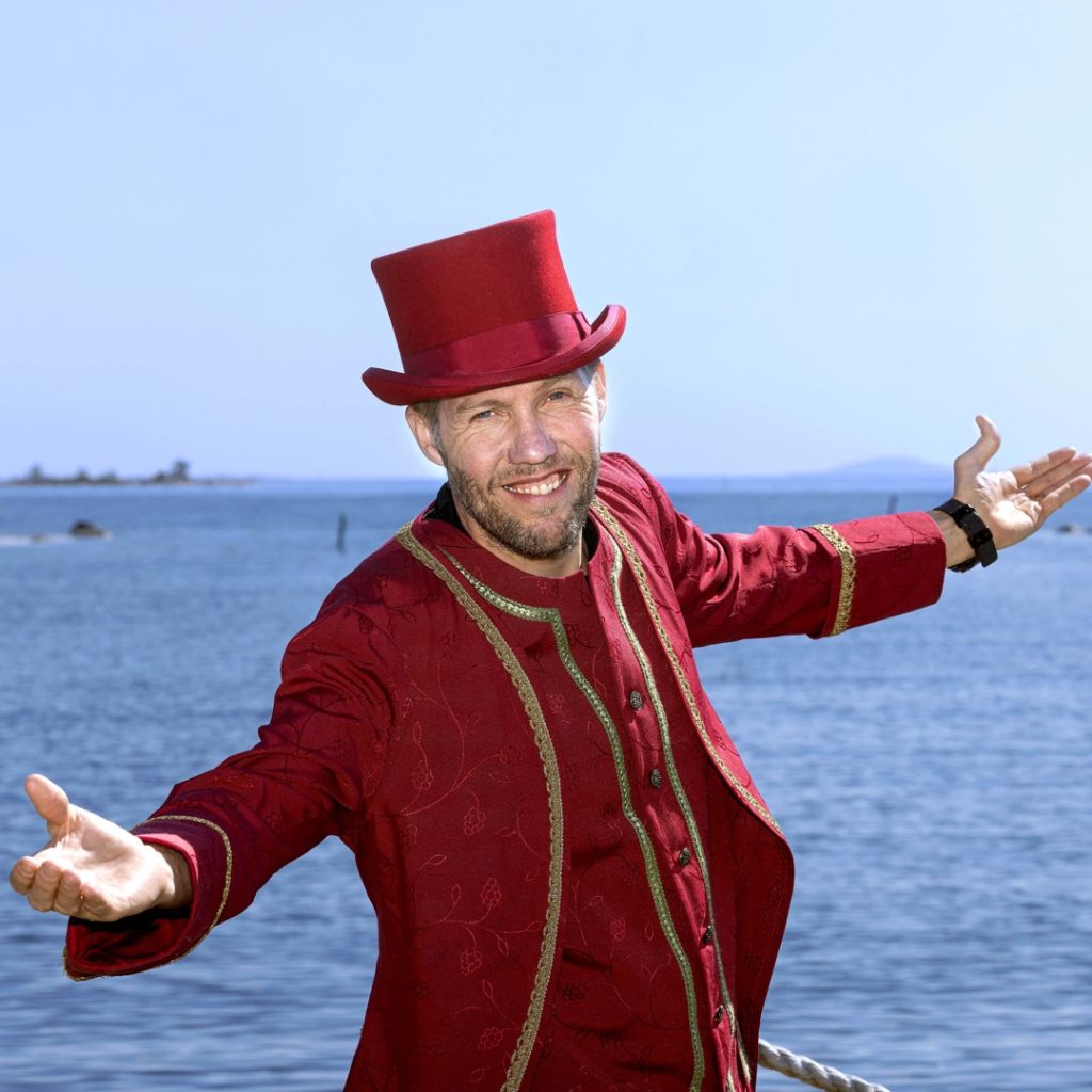 Johan Erlansson poserar framför havet med utsträckta armar, iklädd röd frack och hatt.